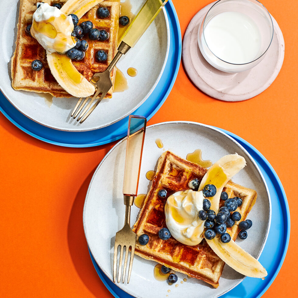 Buttermilk waffle recipe, two waffles with bananas, blueberries and yoghurt, served on white plates with an orange background